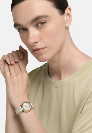 Vrouw met naar achteren gekamd haar, gekleed in een beige blouse, die een horloge met een witte leren band, een gouden kast en kristallen uurmarkeringen toont.