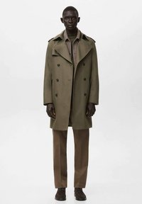 Gabardina de doble botonadura de color verde oliva con hombreras, detalles de botones y bolsillos laterales, llevada sobre una blusa beige y pantalones marrones.