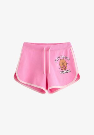Ung person med briller iført grå tank top og lyserøde shorts med tegneserie capybara med donut og tekst 'relaxation', stående indendørs.