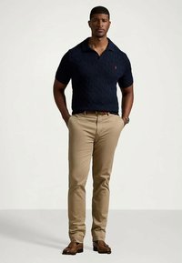 Mężczyzna stoi z rękami w kieszeniach, ubrany w granatowy, krótki sweter polo, beżowe spodnie, brązowy pasek i brązowe mokasyny, na jednolitym tle.