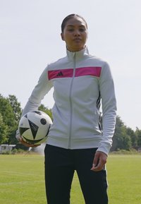 Biała sportowa marynarka z różowym paskiem i czarnymi akcentami. Czarne spodnie. Trzyma czarno-biało wzorzysty piłkę nożną. Teren na świeżym powietrzu.