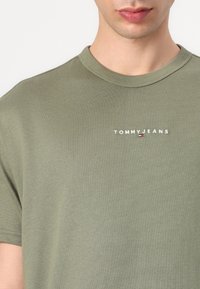 Alyvų žalių medvilnės marškinėlių su apvalia iškirpte, su baltu siuvinėtu "TOMMY JEANS" logotipu ir mažai spalvota emblema kairėje pusėje.