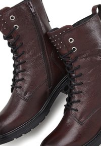 Stivaletti in pelle marrone con superficie texurizzata, dotati di zip laterale, lacci e borchiette decorative sulla parte superiore. Suola in gomma nera.