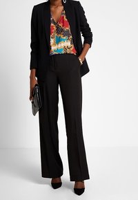 Svart kavaj över en flerfärgad mönstrad blus och svarta vida byxor. Tillbehören inkluderar en svart kuvertväska och spetståskor.