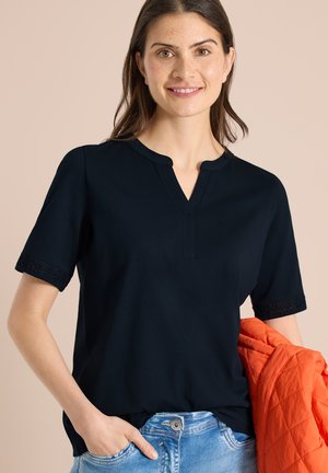 Femme portant un chemisier noir à manches courtes avec des poignets en dentelle, un jean bleu, et tenant une veste matelassée orange sur un fond beige.