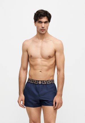 Jeune homme aux cheveux courts et foncés, torse nu, portant un boxer bleu marine avec une ceinture noire et dorée à motif.