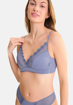 Femme portant un ensemble assorti soutien-gorge en dentelle violet clair et sous-vêtements avec des motifs floraux, posant avec une main près de son épaule.
