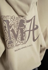 Haut à capuche beige présentant un grand graphique marron, combinant des lettres stylisées « M » et « A » avec des motifs complexes, logo au-dessus de l'ourlet.