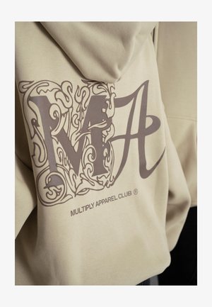 Felpa beige con un grande graphic marrone, che combina le lettere stilizzate "M" e "A" con disegni intricati, logo sopra l'orlo.