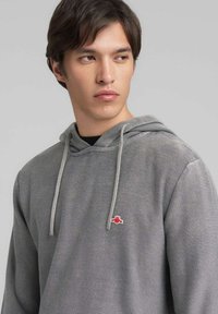 Sudadera gris texturizada con capucha ajustable mediante cordón y un pequeño logo rojo en el pecho, presenta un ajuste relajado y mangas largas.
