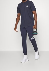 Námořnické modré sportovní tričko a odpovídající tepláky, oboje s bílými akcenty loga Adidas. Bílé tenisky a kettlebell doplňují celý outfit.