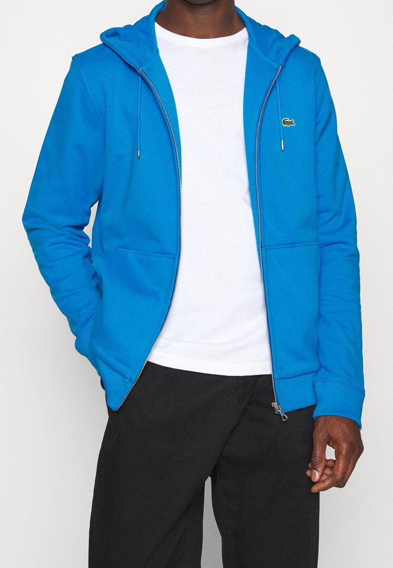 Homme portant un sweat à capuche bleu vif zippé par-dessus un t-shirt blanc et un pantalon noir, debout avec une main dans la poche et l'autre détendue.