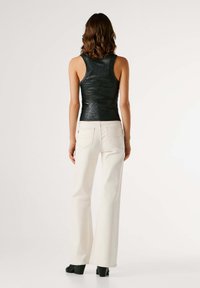 Μαύρο αμάνικο τοπ με γυαλιστερή υφή, συνδυασμένο με λευκά τζιν με φουντωτή γραμμή. Μαύρα παπούτσια με χοντρό τακούνι. Το μοντέλο στέκεται με την πλάτη γυρισμένη προς τον θεατή.