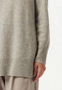 Ljusgrå stickad tröja med ribbad nederkant och ärmslut, som visar en mjuk textur. Fokuset ligger på tyget och detaljen på ärmarna.