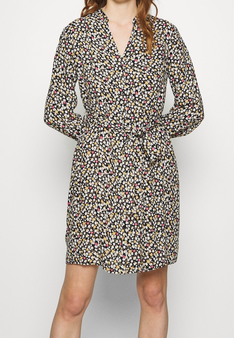 Femme portant une robe noire à manches longues avec un imprimé floral petit en blanc, jaune et rose, et une ceinture nouée à la taille.