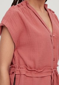 Gros plan sur une personne portant une robe sans manches rose poussiéreuse à boutons texturée avec une taille à cordon et des cheveux tressés.