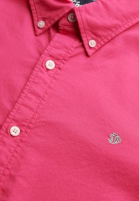 Różowa bawełniana koszula z guzikami, z pointed kołnierzykiem, białymi guzikami i małym zielonym haftowanym logo na piersiach.