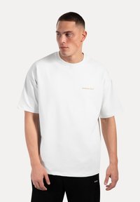 Weißes Baumwoll-T-Shirt mit kurzen Ärmeln, rundem Hals und leicht beigem gesticktem Markenlogo auf der linken Brust. Glatte Textur.