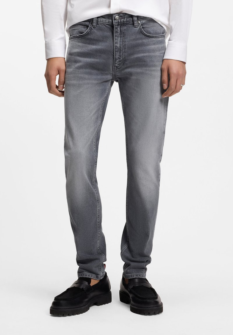 Jeans de mezclilla gris ajustados con acabado descolorido, diseño de cinco bolsillos y costuras en contraste, combinados con zapatos negros sin cordones.