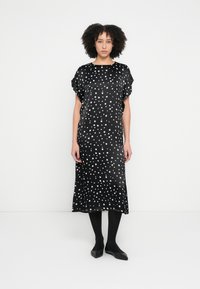 Schwarzes Satinkleid mit weißen Polka-Dots, kurzen Flatterärmeln und lockerem Schnitt, reicht bis zur Mitte der Wade, kombiniert mit schwarzen Strumpfhosen und Schuhen.
