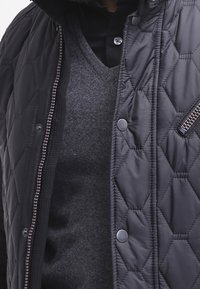 Czarna pikowana kurtka z heksagonalnym wzorem, srebrnym zamkiem błyskawicznym i zatrzaskami. Noszona na szarym swetrze z dekoltem w serek. Widoczna miękka faktura.