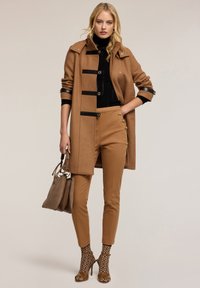Bruine wollen jas met zwarte accenten, aangename zwarte trui, tan kleurige broek met gouden knopen, visnetpatroon sokken en naakte hoge hakken.