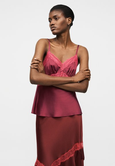 Γυναίκα με σταυρωμένα χέρια, φορώντας αμάνικη μαρουνί σατέν μπλούζα με δαντελένιο τελείωμα και αντίστοιχη φούστα, μπροστά σε λευκό, απλό φόντο.