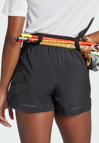 Shorts de sport noirs avec une texture lisse, dotés d'accents réfléchissants et d'une taille sécurisée tenant des poteaux d'entraînement lumineux.