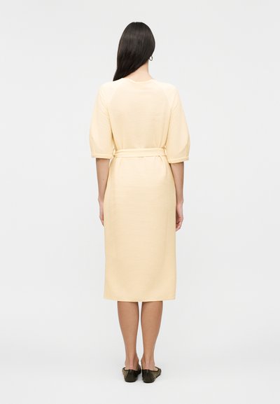 Γυναίκα με μακριά σκούρα μαλλιά, που φοράει ένα ανοιχτό κίτρινο midi φόρεμα με μανίκια μέχρι τον αγκώνα και ζώνη στη μέση, στέκεται γυρισμένη με την πλάτη, σε απλό λευκό φόντο.