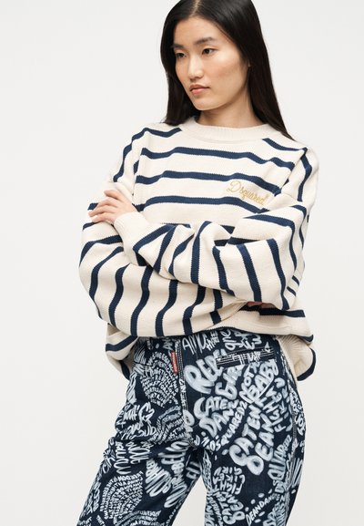 Ριγέ μπεζ πουλόβερ με ναυτικές ρίγες και χρυσό λογότυπο· συνδυασμένο με μαύρο τζιν παντελόνι με λευκά γραφικά κείμενα.