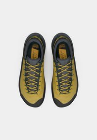 Botas de senderismo La Sportiva en amarillo y gris con suelas negras y cordones verdes y amarillos, mostradas desde arriba sobre un fondo blanco.