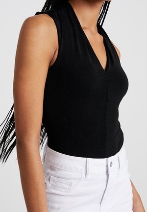 Zwart mouwloos V-hals topje gedragen met een witte broek met hoge taille, zijaanzicht van het bovenlichaam en gevlochten haar.