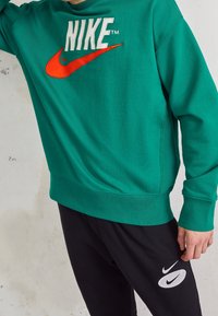 Person som bär en grön Nike-tröja och svarta Nike-sweatpants med synliga logotyper, stående mot en ljus väggbakgrund.