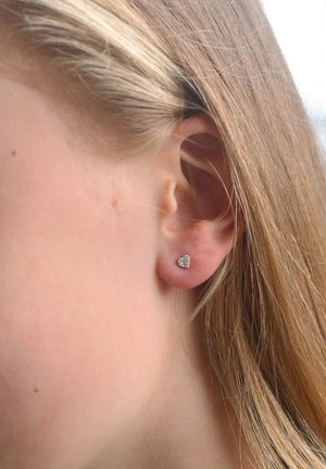 Boucles d'oreilles en argent en forme de cœur, fixées sur une petite base métallique, portées sur le lobe d'une oreille à la peau claire avec des cheveux raides et châtain clair.
