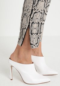 Pantalon imprimé serpent longueur cheville avec fermeture éclair latérale, associé à des mules blanches à talons hauts et bouts pointus sur un fond clair.