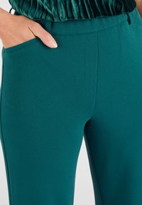 Teal broek met een gladde textuur, voorzien van zijzakken en een aansluitende tailleband. De stof heeft een lichte stretch voor comfort.
