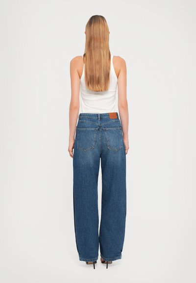 Jeans en denim à jambes larges avec un délavage bleu moyen, dotés d'une étiquette en cuir à la taille et de deux poches arrière. Texture lisse, design minimal.