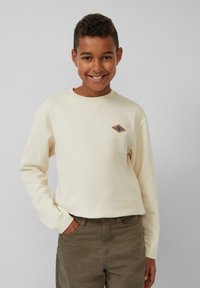 Crème sweatshirt met lange mouwen en een diamantvormig geborduurd detail in blauw en oranje op de borst. Gepaard met olijfgroene broeken.