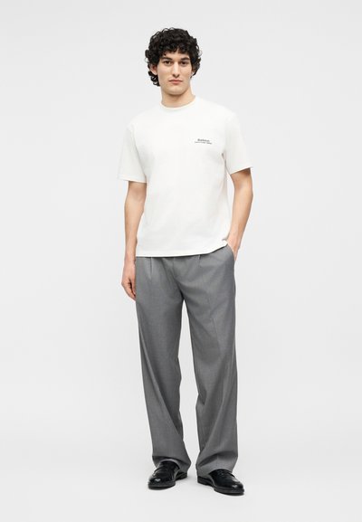 Νέος άνδρας με σγουρά μαλλιά, φοράει λευκό κοντομάνικο πουκάμισο, γκρι παντελόνι και μαύρα παπούτσια, στέκεται με το ένα χέρι στην τσέπη.