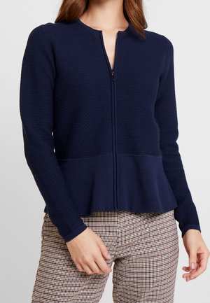 Femme portant un cardigan zippé texturé bleu marine avec un ourlet à volants et un pantalon à carreaux, mains détendues sur les côtés.