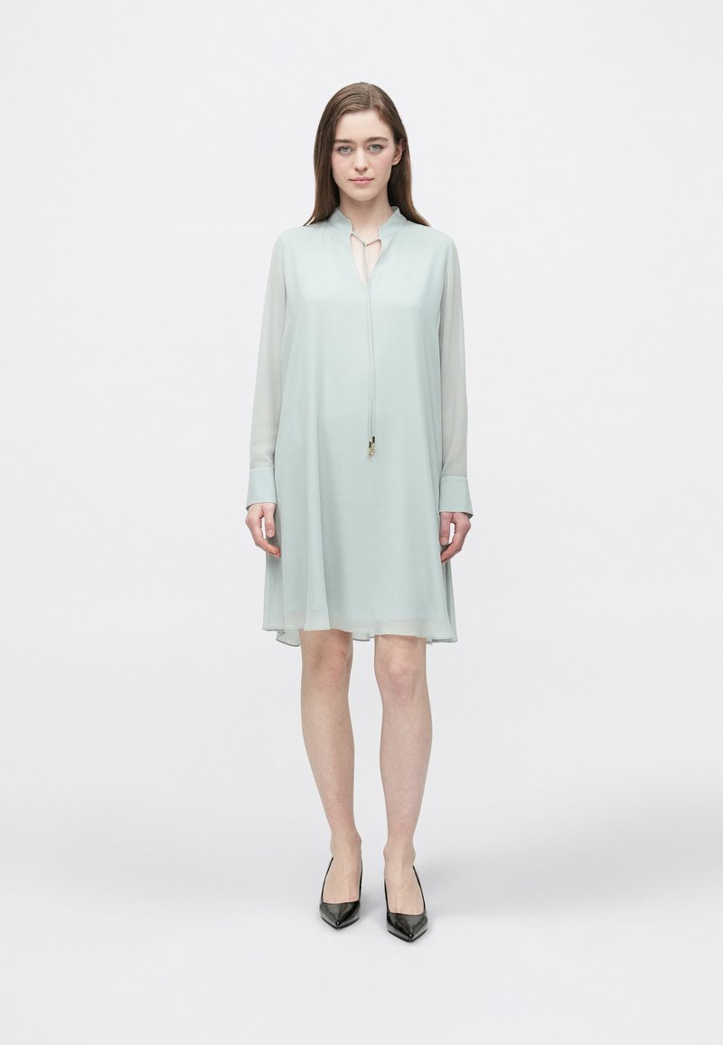 Hellgrünes, knielanges Kleid mit Kragen, langen, durchscheinenden Ärmeln und einer Frontschleife. Kombiniert mit schwarzen, spitz zulaufenden Absatzschuhen. Glatte Textur.