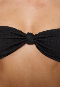 Top de bikini bandeau preto com um padrão nervurado texturizado, apresentando um detalhe central em nó. Design liso e ajustado.