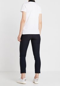 Person som står med ryggen mot kameran iförd en vit kortärmad skjorta, svarta smala byxor och vita sneakers, på en enfärgad vit bakgrund.