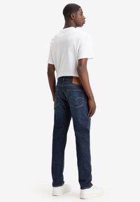 Jeans de color azul oscuro con un corte recto, fabricados en tela de mezclilla, con costuras en contraste y una etiqueta de cuero marrón en la parte trasera de la cintura. Combinados con una camiseta blanca.