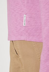 Vaaleanpunainen lyhythihainen paita pehmeästä kankaasta; siinä on sivutagi, jossa lukee "Marc O'Polo Denim." Yhdistetty havainnollistaviin beigeihin housuihin, joissa näkyy tasku.