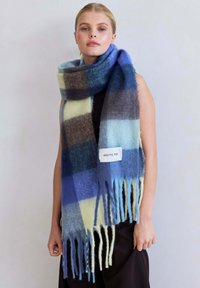 Overmaatse wollen sjaal met een blauw, groen en bruin ruitpatroon en franjes aan de randen. Etiket leest "ARCTIC FOX." Zachte textuur.