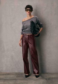 Grå off-shoulder sweater, bordeaux læder bukser med vide ben, sort taske og hæle; solbriller fuldender looket mod en grå baggrund.