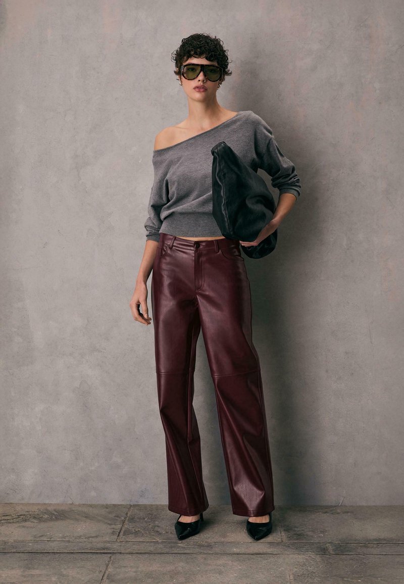 Grå off-shoulder sweater, bordeaux læder bukser med vide ben, sort taske og hæle; solbriller fuldender looket mod en grå baggrund.