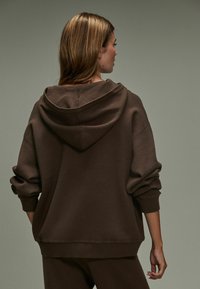 Felpa con cappuccio marrone dal taglio relaxed, con spalle scese e polsini a costine, presentata su uno sfondo neutro.