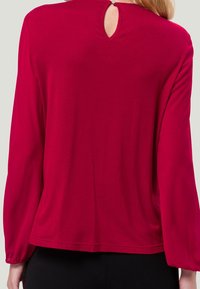 Blouse rouge à manches longues avec un détail en keyhole au col à l'arrière. Texture lisse et tissu doux. Manches élastiques et coupe décontractée.